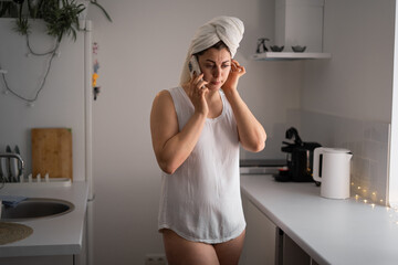 Woman with towel on head talking on mobile phone standing in morning kitchen at home. Female having...