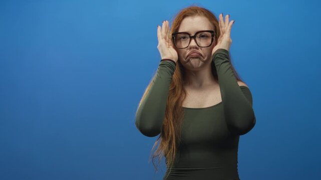 Redhead woman covering face with hands and wearing oversized glasses against a blue studio backdrop; playful coy.