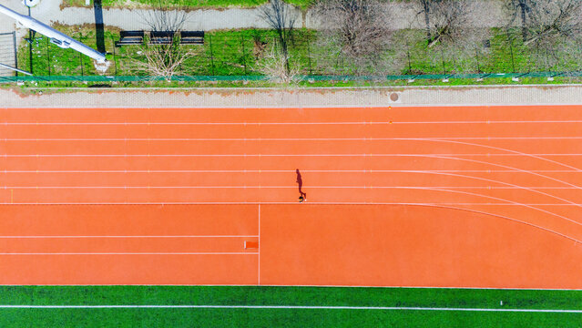 Aerial view of a lone figure strides across the vibrant orange athletic track, framed by lush green fields, capturing a moment of solitary motion, Novi Sad, Vojvodina, Serbia.