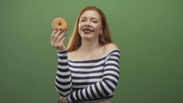 Redhead woman holding a glazed donut with finger to lips shushing in studio wearing striped off shoulder top presenting the treat; playful tease.