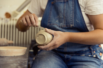 Close-up of a potter in denim overalls trimming the base of a clay bowl with a metal tool in a...