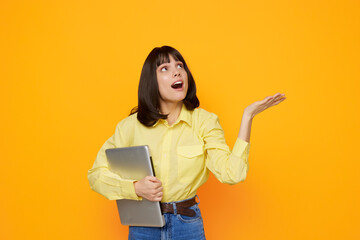 A cheerful young woman stands against a vibrant orange wall, holding a laptop and looking up with wonder, as if discovering a new idea or creative spark. © SHOTPRIME STUDIO