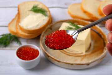 Close-up of red caviar on a spoon. Red caviar in a white bowl, butter, wheat bread and a sprig of dill on a ceramic plate in the background. Ingredients for a sandwich. Superfood, healthy eating