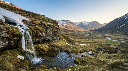 Naklejka premium Waterfall flows from melting snow on mountain slope in spring valley. Scenic landscape with river winding through grassy field during thaw. Nature travel destination in wilderness.