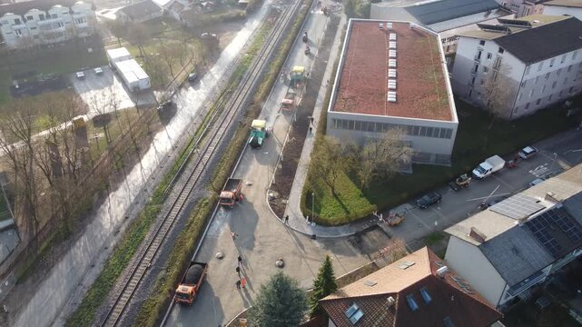 An aerial view of road paving in Siska, Ljubljana, featuring heavy machinery and trucks laying fresh asphalt alongside railway tracks, with the St. Francis of Assisi Church visible in the background.