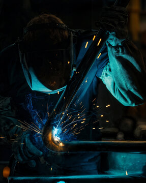 Professional welder wearing protective mask works on metal pipe with bright blue sparks