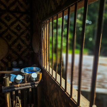 Warm sunlight entering through a wooden window illuminating a rustic bamboo kitchen interior with traditional cooking utensils. The peaceful scene captures natural light and rural atmosphere.