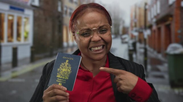 Woman smiling broadly holds canada passport and points finger to passport on rainy street; pride achievement.