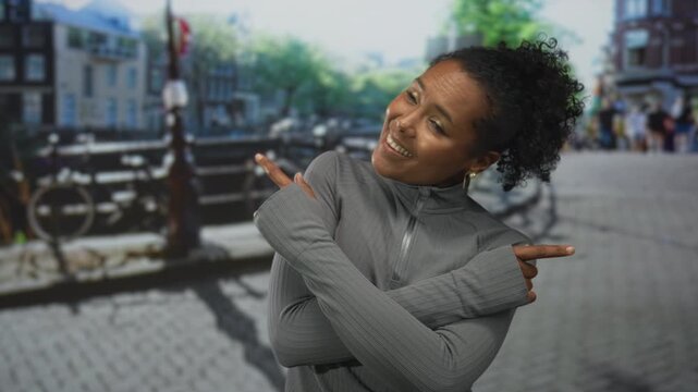 Young african american woman in grey jacket smiles and points finger to building on city street; confidence.