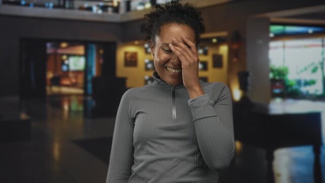 Young african american woman laughing and covering her face with her hand in a hotel lobby near seating area and reception desk; bashful happiness.