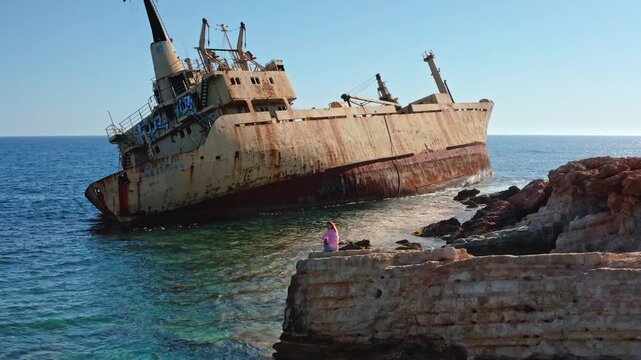Young Caucasian woman sitting on coastal rocks, looking at the rusty Edro III shipwreck partially submerged in the clear turquoise waters of the Mediterranean Sea near Paphos, Cyprus