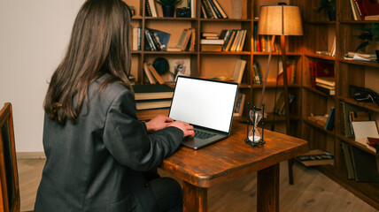 Woman working on laptop in a cozy library with bookshelves and hourglass on desk © shine.graphics