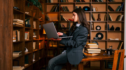 Young woman in a suit working on a laptop in a cozy library with bookshelves © shine.graphics