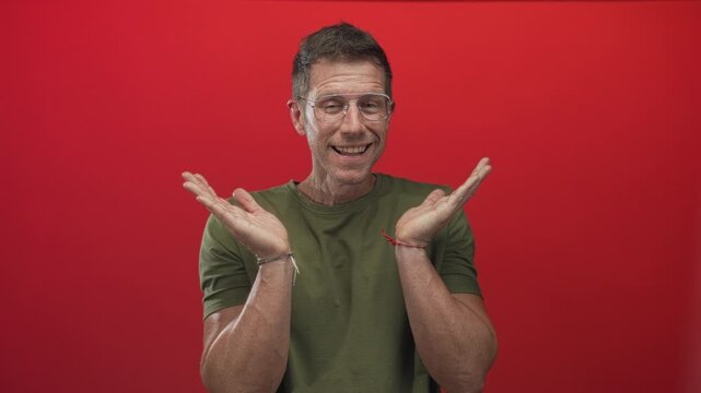 Man covering face with hands in red studio wearing olive green t shirt and clear glasses; embarrassment.