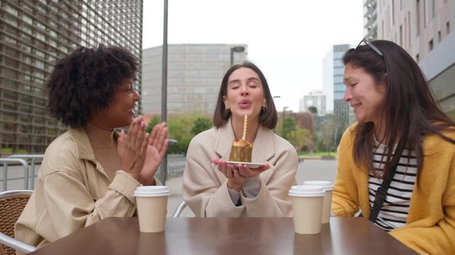 Diverse young women celebrating a birthday as a friend blows out a candle on a mini cake at an outdoor cafe