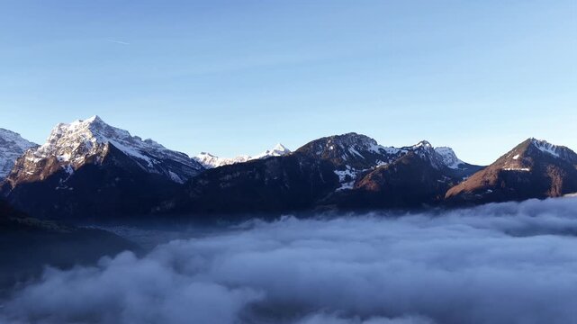 Snowy Swiss Alps rise above a dramatic sea of fog over Walensee Lake at sunrise. Serene alpine landscape with glowing peaks and cloud inversion, ideal for travel, nature, and climate.