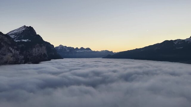 Soft sunrise light spreads across a vast sea of fog covering Walensee valley in the Swiss Alps. Peaceful alpine winter scene with dramatic silhouettes and tranquil morning atmosphere.