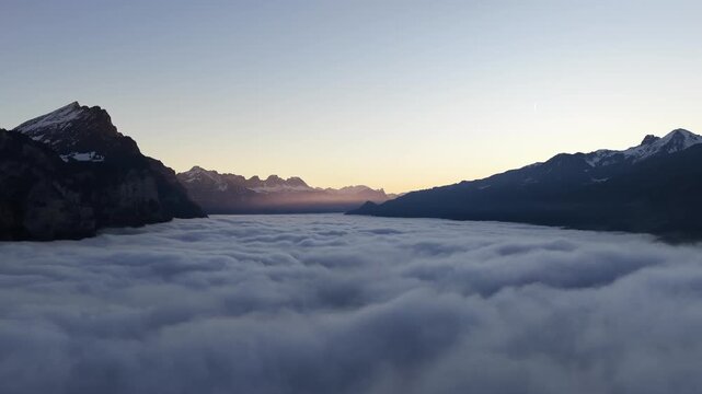 Golden sunrise light touches the alpine horizon above Walensee, Switzerland, while mountain silhouettes emerge from a vast sea of fog covering the quiet valley below.
