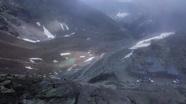 Drone footage captures the dramatic alpine terrain of Sach Pass and Pangi Valley, featuring rocky slopes, snow patches, glacial pools, and misty mountain peaks in the Indian Himalayas.