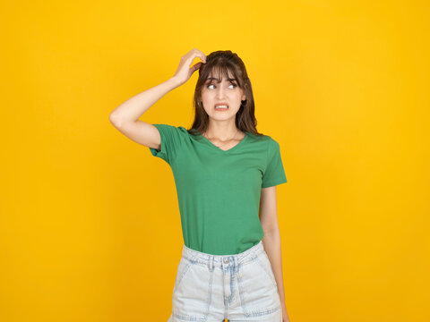 Confused young woman scratching her head while making an uncertain facial expression, standing against a vibrant yellow studio background with wide copy space, wearing a green t shirt.