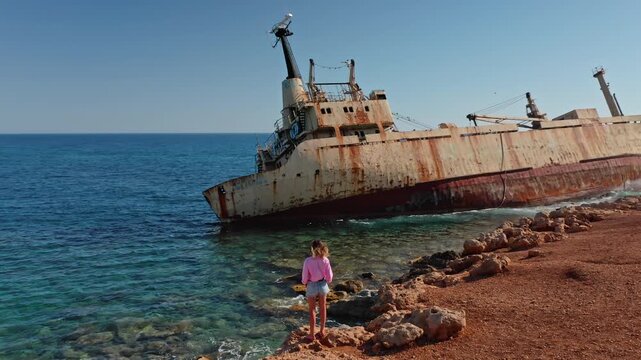 Woman standing on the rocky seashore admiring the iconic rust-covered Edro III Shipwreck grounded on the coast of Paphos, Cyprus, with clear blue Mediterranean sea and sky