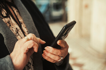 Close up of senior woman hands using a smartphone while walking in a city street with empty space for text or promo.