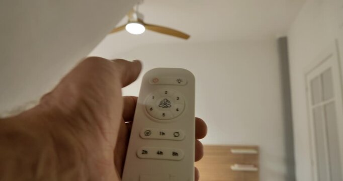 Fan Control: A hand expertly operates a remote control, managing the speed and light of a ceiling fan in a modern interior, symbolizing home comfort and ease.