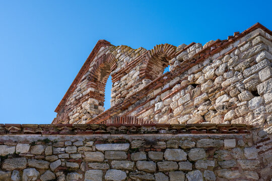 Low-angle view of the Church of Saint Sophia ruins in Nesebar, Bulgaria, highlighting the ancient stone walls, brick detailing, and a high arched window against a clear blue sky.
