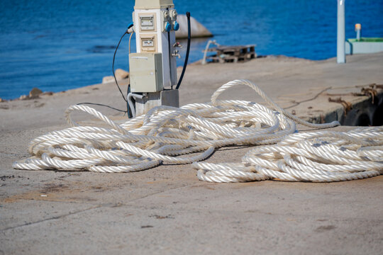 Coiled white nautical rope lying on a concrete pier with a power pedestal and the blue sea in the background.