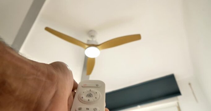 Ceiling Fan Control: A hand aims a remote at a sleek ceiling fan with three blades, the soft glow of the integrated light fixture illuminating the space. A modern approach to staying cool