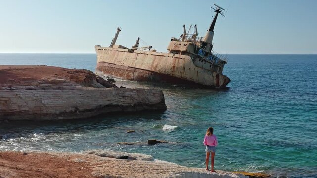 Young girl and others on rocky Paphos coastline watching the stranded Edro III cargo ship against clear blue Mediterranean waters and a bright summer sky, travel and nature clash