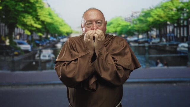 Man monk covers mouth with hands in street by a canal, brown robe visible and eyes closed in a prayerful posture; contemplation devotion.