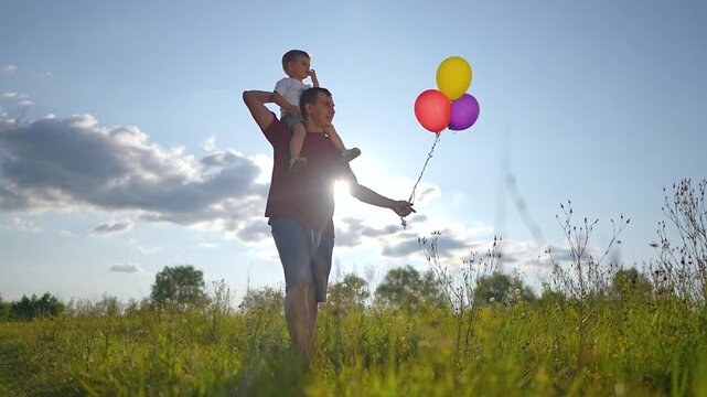 Father carries child on shoulders with colorful balloons. Dad walks in meadow holding balloon strings. Happy son rides on father outdoors. Parent and child enjoy sunny day together in field.