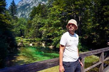 Fototapeta premium man in bucket hat standing by the crystal clear turquoise river in Slovenian Alps