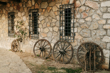 Fototapeta premium Old wooden wagon wheels leaning against rustic stone wall with iron window bars. Traditional countryside architecture concept representing rural heritage and historical transport