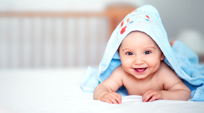 Happy smiling baby boy lying on bed covered with a blue hooded towel. Adorable infant after bath time in a bright nursery setting with copy space for parenting and childcare concepts