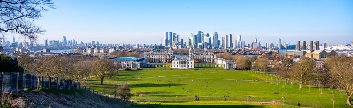 Visitors enjoy a sunny day in Greenwich Park, surrounded by greenery. The historic buildings contrast with the modern London skyline in the background, creating a beautiful scene.
