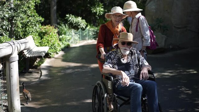 Mature woman pushes her father in a wheelchair while her mother walks beside them at the zoo in San Diego, having a fun day out together.