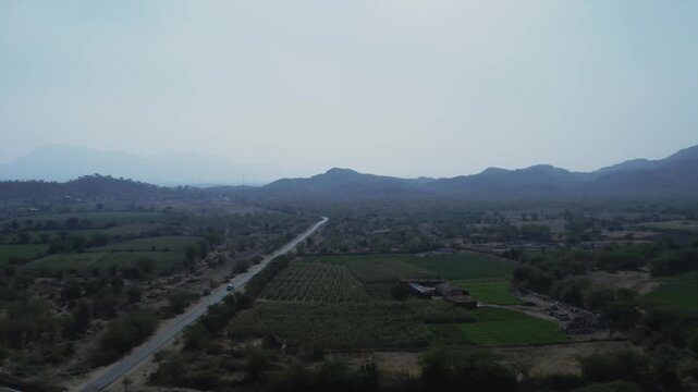 A single vehicle travels along a winding paved highway cutting through cultivated farmland and rural terrain in Rohida, Rajasthan, with distant mountain ridges under hazy atmospheric light.