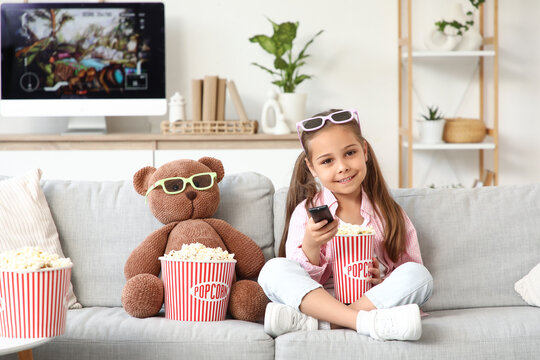 Cute happy girl in 3d glasses with teddy bear, popcorn and TV remote control watching cartoons on sofa at home