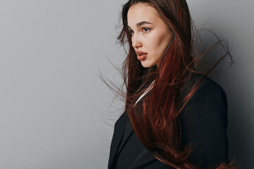 Young woman with long brown hair wearing black jacket stands against gray background with hair blowing, looking to side with serious expression and natural makeup. © SHOTPRIME STUDIO