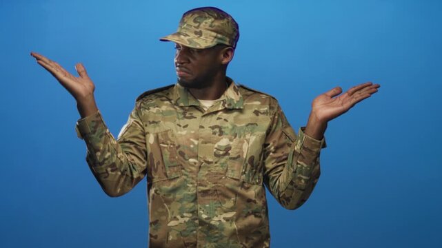 Soldier man in camouflage uniform raises both palms up in a perplexed shrug in a blue studio; confusion.