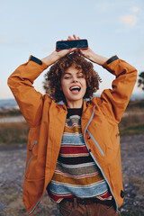 Happy woman with curly hair wearing rainbow sweater and orange jacket smiles holding smartphone...