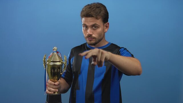 Young hispanic man holding gold trophy points finger toward camera in blue studio after win; victory pride.