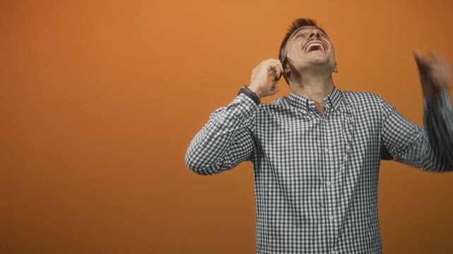Man with mustache in checkered shirt holding smartphone to ear with hand, smiling, leaning forward and raising fist in studio; joy celebration success.
