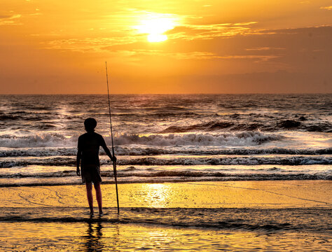 Silhouette of a teenage boy surf fishing at sunrise at the beach with waves in background
