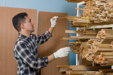 Asian male professional carpenter carefully pulling thin wooden strip from stacked shelves during...