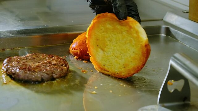 Cook in black rubber gloves carefully flips sliced hamburger bun. Chef flips bun letting aroma of toasted dough fill kitchen. Slow motion