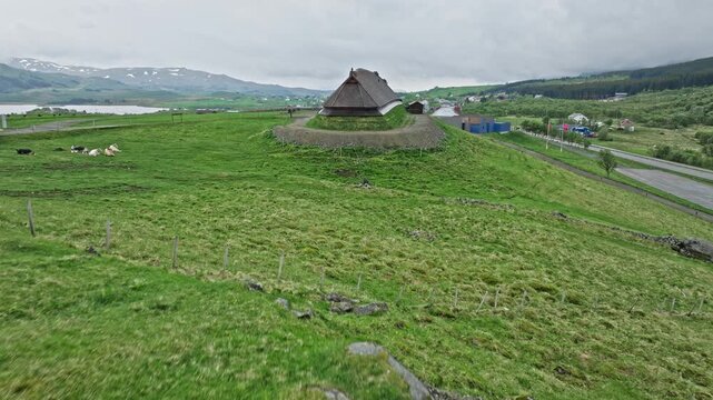 Aerial view of the Lofotr Viking Museum longhouse in Borg, Lofoten Islands, Norway. Historic Viking site surrounded by green fields and rural Norwegian landscape under cloudy sky.