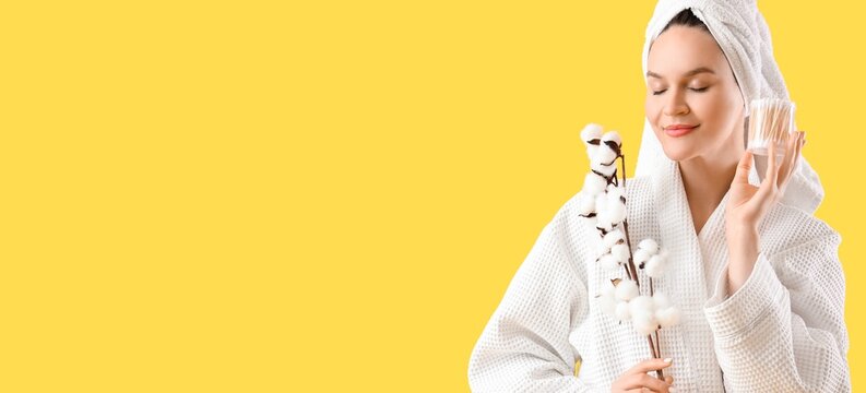 Young woman in bathrobe holding cotton buds and branch on yellow background with space for text